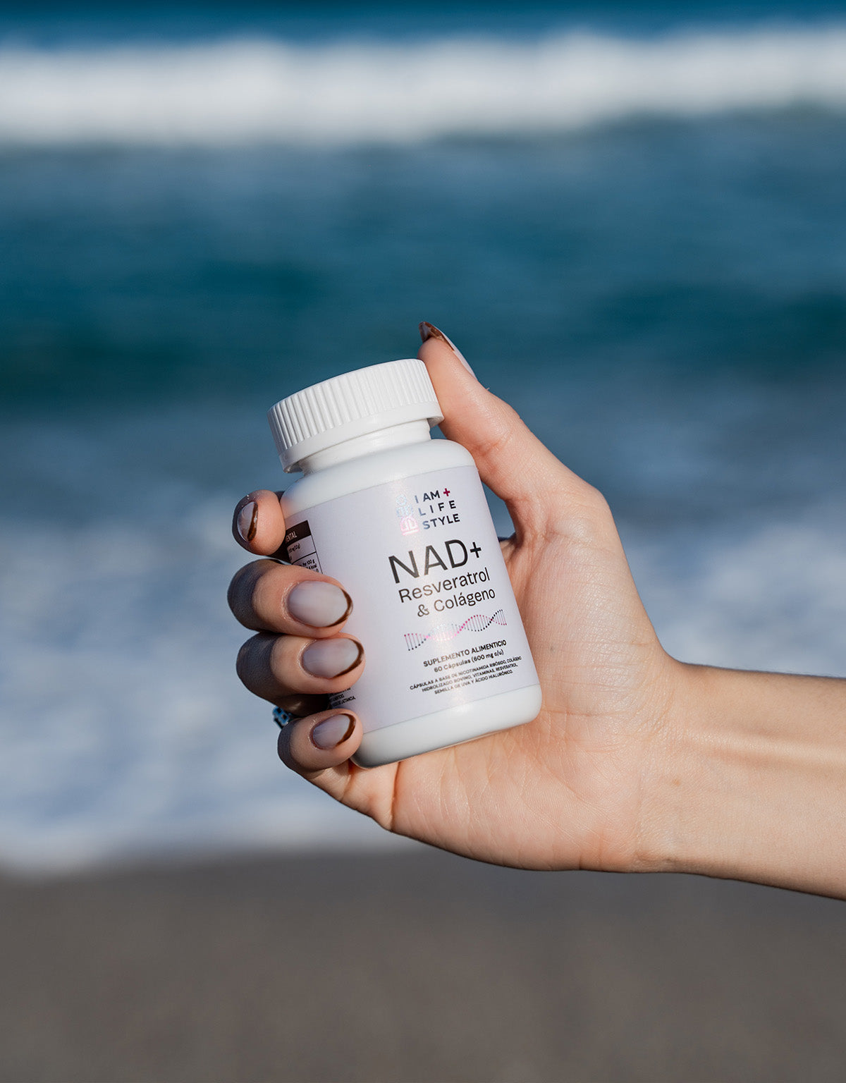 Primer plano de una mano sosteniendo un frasco blanco de suplemento IAM+LIFESTYLE NAD+ Resveratrol & Colágeno con el océano azul de fondo.