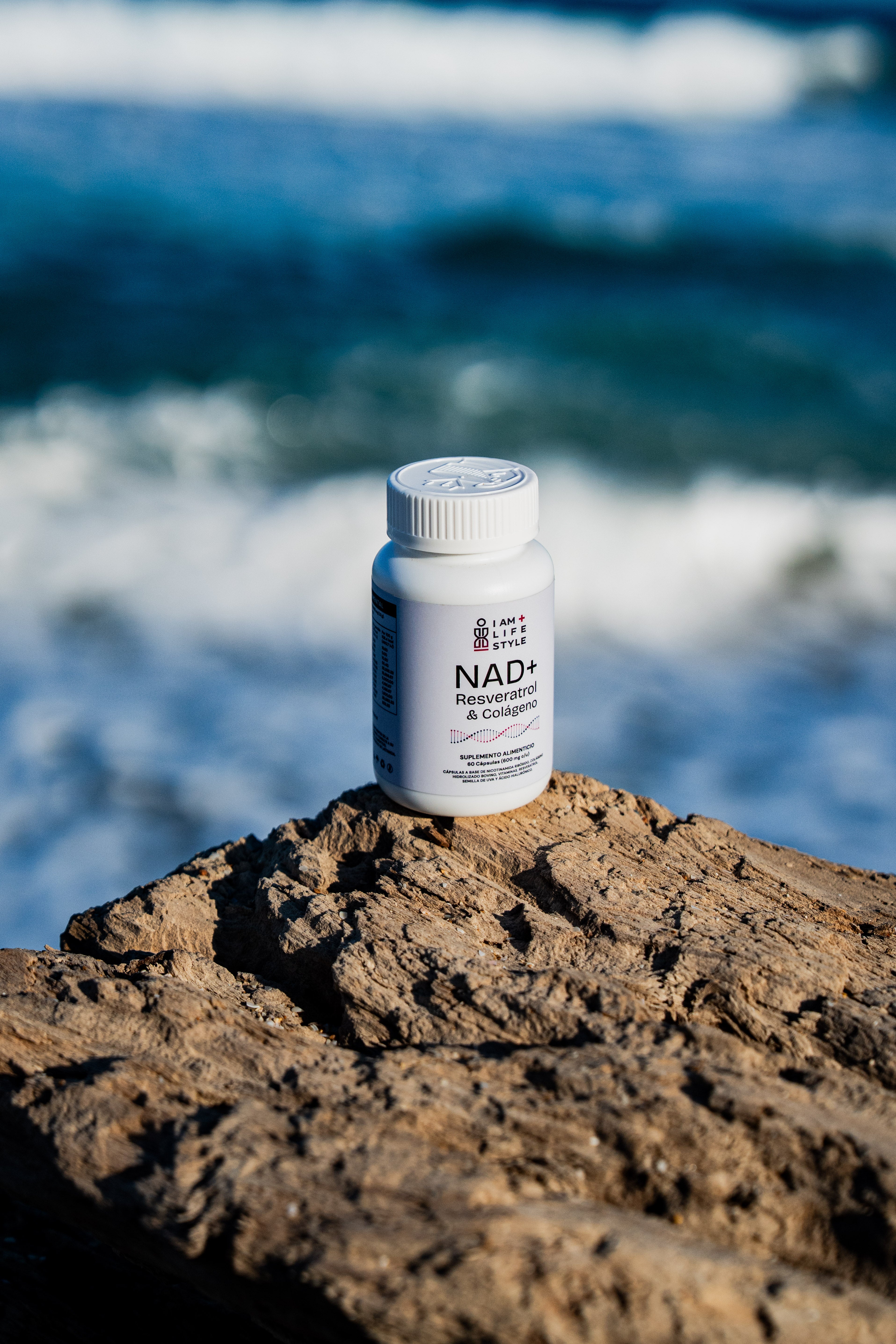Frasco blanco de suplemento IAM+LIFESTYLE NAD+ Resveratrol & Colágeno sobre un tronco de madera rugoso en la orilla del mar, con el océano azul de fondo.