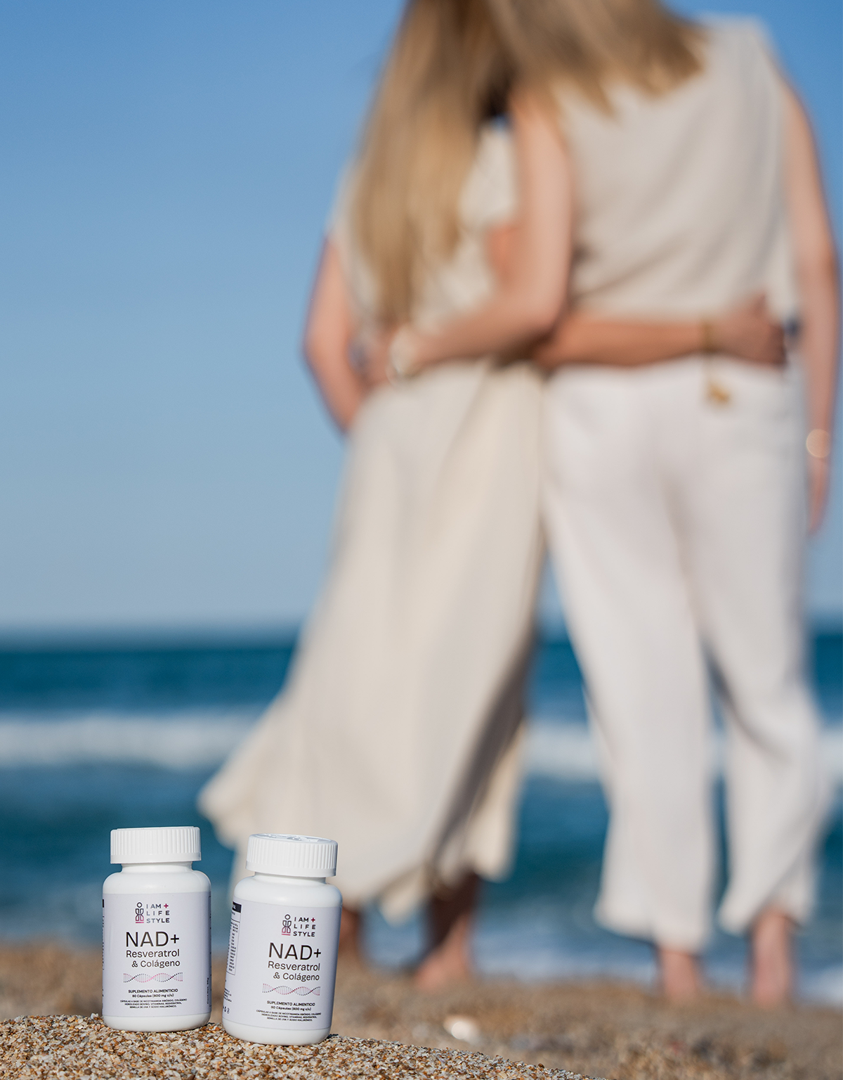 Dos frascos del suplemento NAD+ Resveratrol Colágeno IAM+LIFESTYLE en la arena, con dos mujeres abrazadas con el mar de fondo, simbolizando bienestar y conexión.